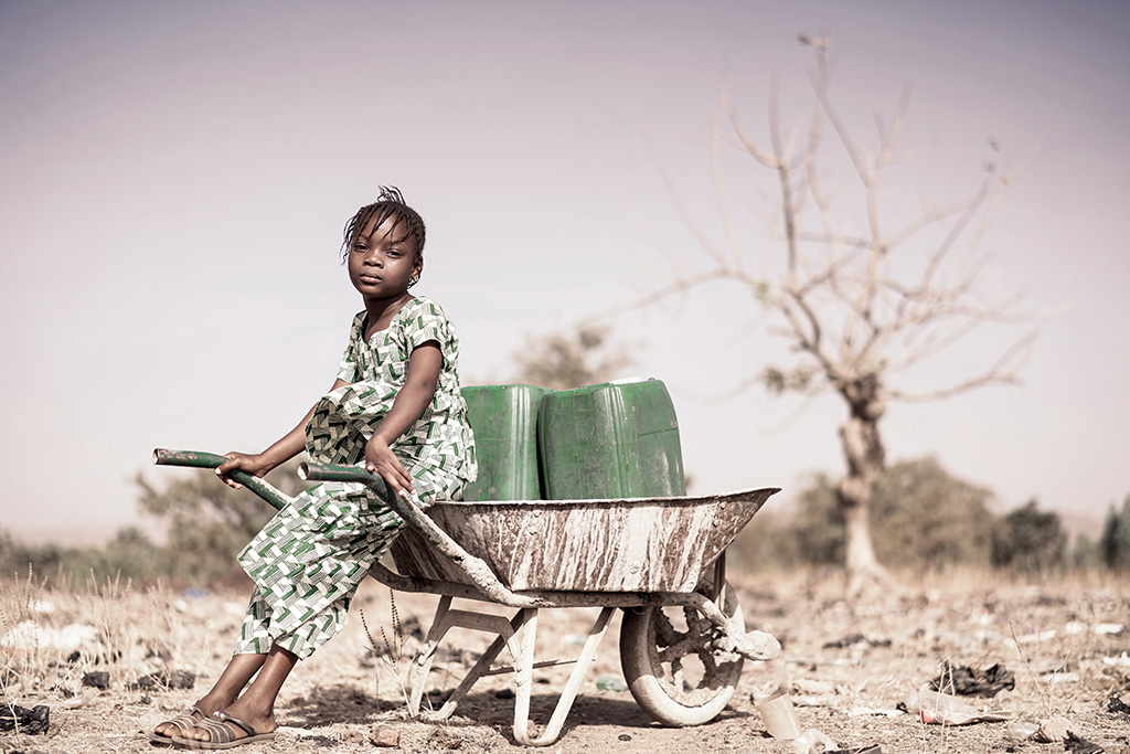 Foto av jente med trillebåre og vannkanne i tørt land.